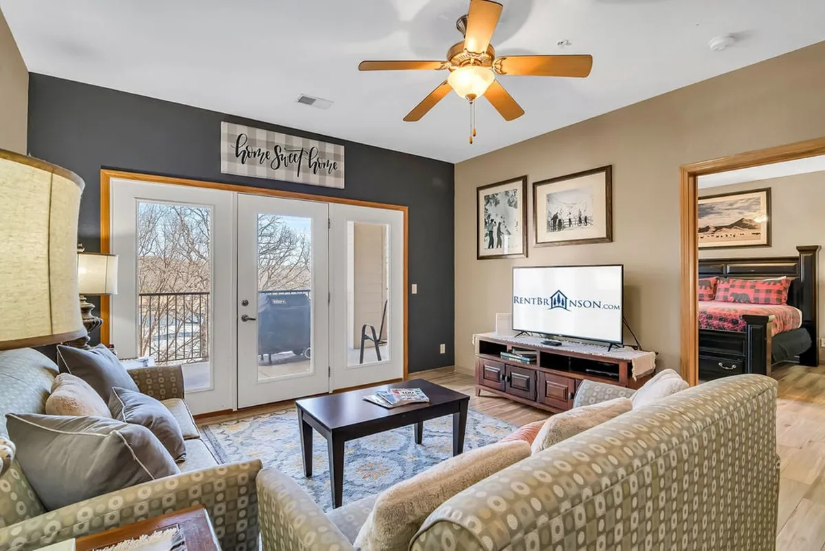 A welcoming living room with a comfortable sofa (which can double as a sleeper), a television, decorative wall art, and large balcony doors letting in sunshine and offering a view outside. The arrangement is ideal for relaxing together after a day of adventure.