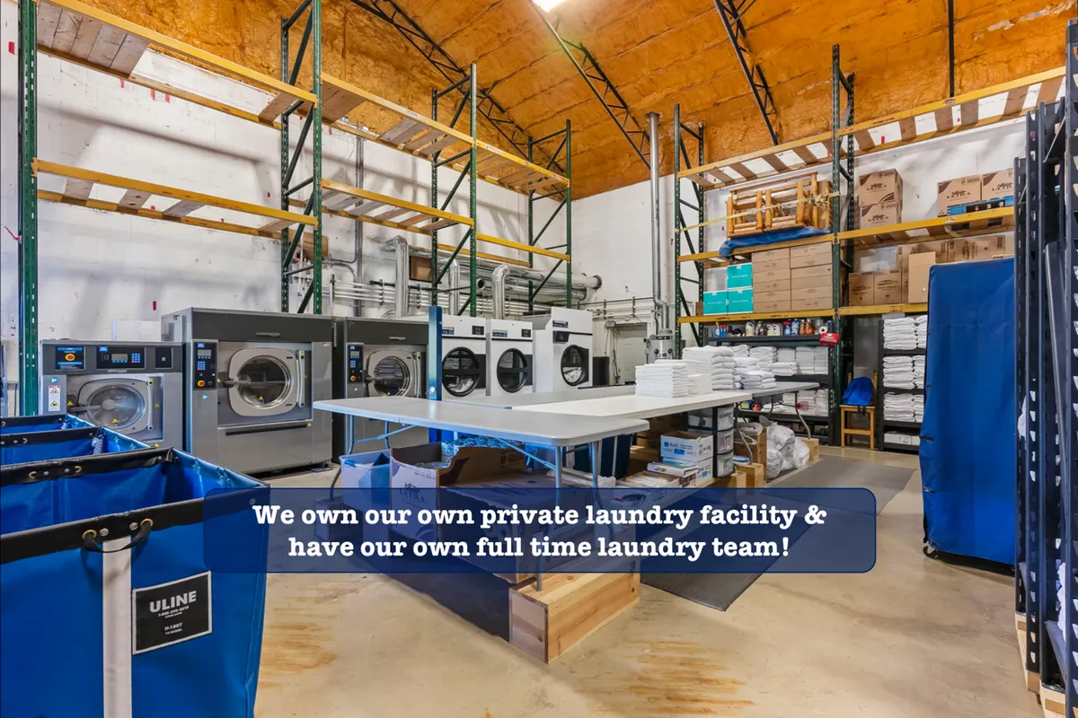 A dedicated laundry space featuring industrial-grade washing machines, a large folding table, shelves stocked with cleaning supplies, and laundry carts. The area is clean, organized, and clearly set up for professional-level cleaning.