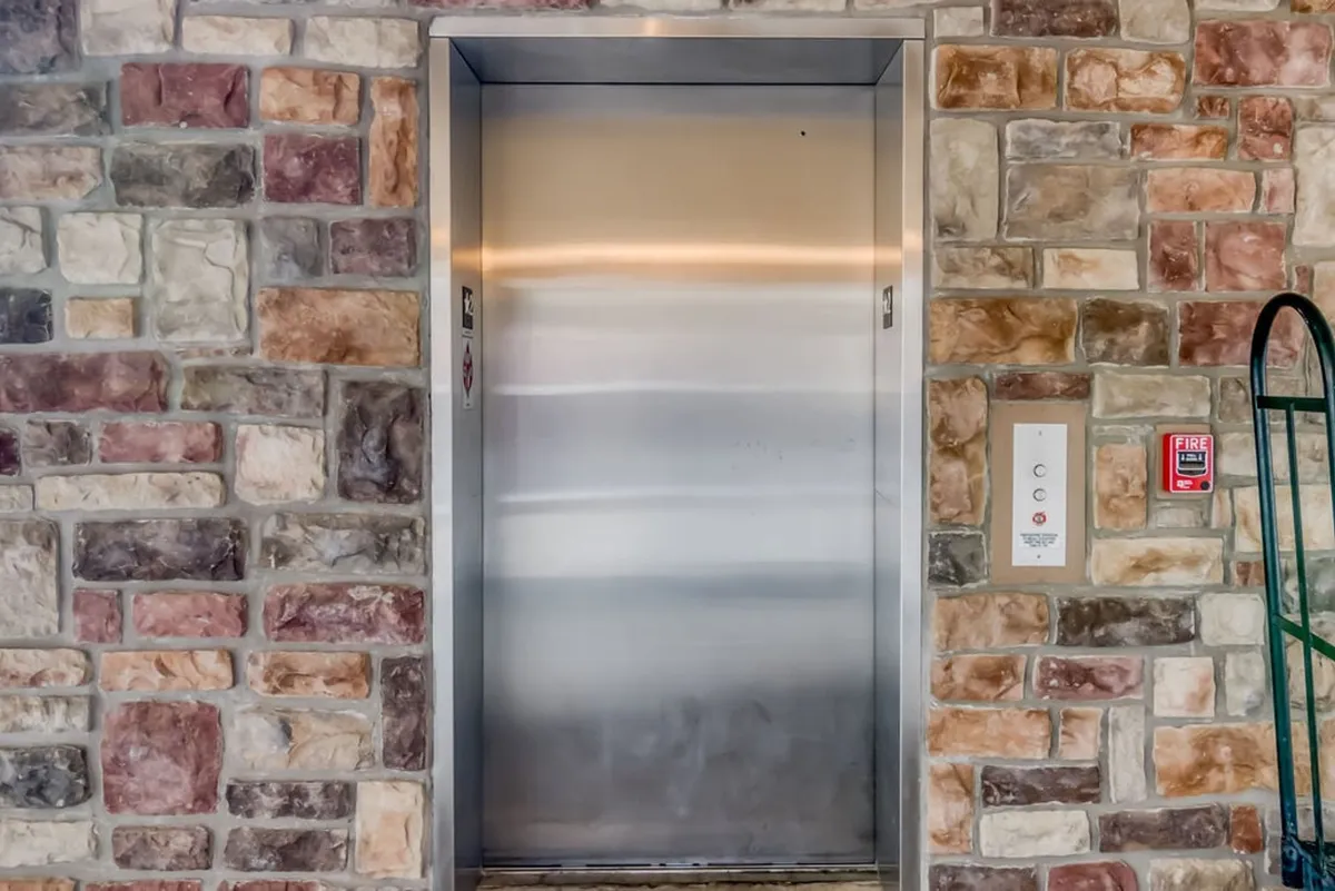 A modern elevator with a stone wall texture and a visible control panel sits in the building’s lobby area. The setting is clean, accessible, and practical, emphasizing effortless movement between floors.