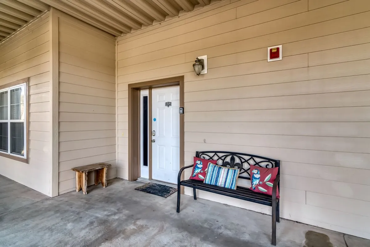 An inviting exterior shot featuring the condo's front door, a welcoming bench adorned with decorative pillows, and the pathway leading up to the entrance. The scene suggests easy access and a friendly atmosphere, with natural daylight hinting at the proximity to outdoor activities.