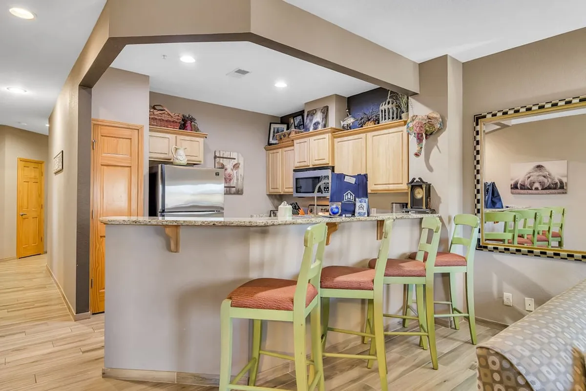An inviting kitchen and living area with a bar counter, wooden cabinets, and stools with green legs. The space is styled for both entertaining and everyday living, with shelves displaying decorative items, and a layout conducive to group gatherings.