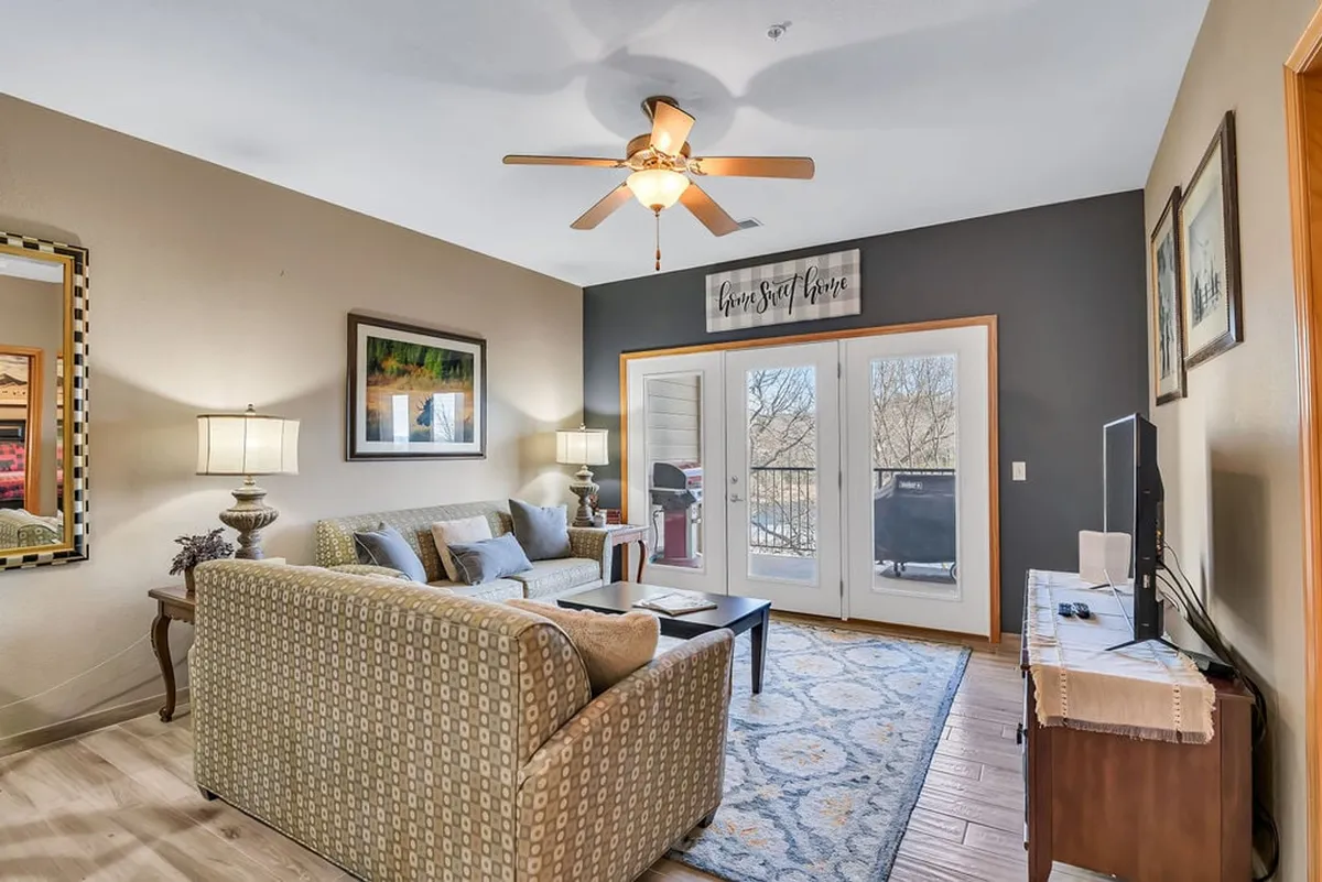The image showcases a warm, inviting living room with plush seating centered around a coffee table, a stylish decorative mirror above the sofa, a TV for entertainment, and sunlit double doors leading out to a private balcony with views of Table Rock Lake.