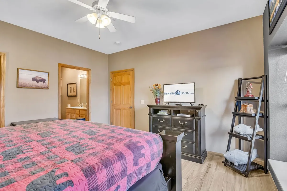 A spacious bedroom featuring a king-size bed with a patterned quilt, a dresser with a television, and a doorway leading to a private bathroom. Sunlight filters through windows, creating a warm and inviting atmosphere.