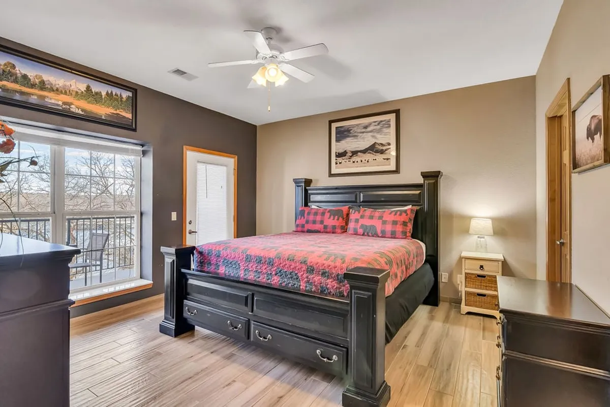 A cozy bedroom featuring a bed with patterned bedding, a large window that opens up to a scenic view, and framed artwork on the wall. The natural light pouring in through the window hints at a connection to the outdoors and the lake just outside.
