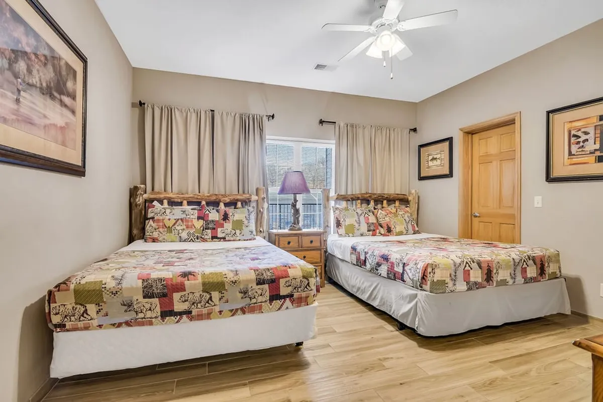 A double queen bedroom with two beds featuring coordinated patterned bedspreads, a window letting in natural light, wooden furniture, and wall art. The room feels spacious and comfortable, perfect for family or friends.